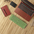 A green wireless foldable keyboard rests on a wooden surface next to a red laptop, notebook, pen, and phone.