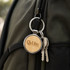 A round wooden keyring with a silver metal ring, attached to a dark green backpack alongside keys.
