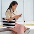 A woman sits outdoors, holding a phone and wearing a black and white striped tote bag with a rope handle.