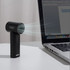 A black handheld fan with a sleek design, positioned next to a laptop on a desk. It features a logo.