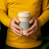A coffee cup made from eco-friendly bamboo fibre, featuring a beige body and grey silicone band. It has a logo.