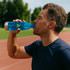 A drink bottle in a bright blue colour, held by a person sipping from it, set against an outdoor track background.