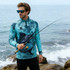 A man wears a blue and teal sublimated 1/4 zip fishing pullover, holding a fishing rod by the sea. It has a logo.