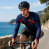 A long sleeve cycling jersey in navy blue and red with a logo, worn by a cyclist on a scenic coastal road.