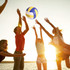 A volleyball with blue and yellow panels is being tossed among several people silhouetted against a sunset.
