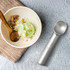 A silver ice cream scoop beside a bowl of green ice cream topped with nuts and oats on a textured surface.