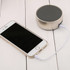 A silver wireless speaker sits next to a smartphone, connected by a white audio cable on a wooden surface.