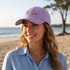A lilac baseball cap with a logo, worn by a smiling woman on a beach, with a scenic ocean view in the background.