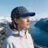 A blue and patterned 5 Panel Custom Peak Cap sits on a woman's head, set against a mountainous landscape with water.