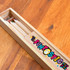 A wooden pencil case containing several coloured pencils, featuring a brand logo and a fun design.