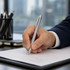 A silver metal pen is held in a hand, writing on a document, with blurred office background items visible.