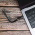 A grey 4-in-1 charging cable is connected to a laptop, resting on a wooden surface. The cable has a logo.