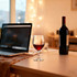 A casual wine glass filled with red wine sits on a table beside a bottle of wine and a laptop.
