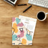 An L-shaped open folder featuring a colourful abstract design, holding a document on a wooden desk with stationery and a coffee cup.