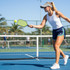 A woman in a white tank top and navy shorts plays pickleball, holding a green paddle and hitting a yellow ball.