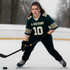 A young woman wearing a green and yellow retro V-neck hockey jersey, poised with a hockey stick on an ice rink.