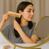 A fine-tooth bamboo small comb in beige held by a person in front of a circular mirror.