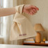 A beige cotton wrist bag is held by a hand, featuring a simple design and a logo.