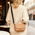A tan polyurethane shoulder bag with a black strap is worn by a person in a cream sweater.
