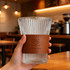 A glass drinkware piece with ribbed design, featuring a brown leather cup sleeve with a logo.