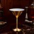 A stainless steel martini goblet with a shiny gold finish, placed on a dark marble surface surrounded by bar tools.