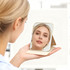 A portable mini mirror in a light gold casing, held by a person, reflecting their face.