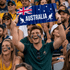 A man holds up a blue cardboard clapper with a kangaroo and Australian flag design, smiling in a cheering crowd.