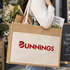 A jute tote bag in natural and beige tones, featuring a logo, being carried with plants and a book inside.