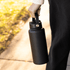 A drink bottle in matte black with a handle, held by a person outdoors. It has a simple design and includes a logo.