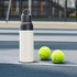 A drink bottle with a black lid and a white body placed next to two green tennis balls on a court surface.