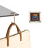 A square, foldable desk hook in metallic colour, with a brown leather-handled bag hanging from it. The hook features a logo.