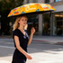 A small, brightly patterned yellow umbrella held by a woman walking on a city street. The umbrella features colourful designs and has a logo.