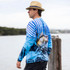 A man wearing a long sleeve blue polyester fishing tee with a fish design, standing by the water with a straw hat.