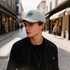 A grey PU leather baseball cap with a logo, worn by a young man in a stylish urban setting.
