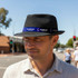 A black mesh top hat with a blue and black branded band, worn by a man in a sunny street setting.