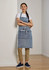 A denim-coloured apron with pockets, worn by a model standing against a wall, paired with a white top and tan trousers.