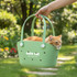 A green pet carrier with handles holds an orange cat, set against a garden background with flowers.
