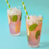 Two tall glasses of a colourful drink with ice, garnished with mint leaves and lemon, featuring green and white striped paper straws.