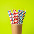 A cup filled with red and white striped and blue and white striped disposable paper straws against a bright green background.