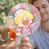 A transparent PVC hand fan featuring a cartoon character with blonde hair and a playful expression, held by a person.