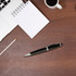 A black metal pen with a sleek design sits on a wooden desk alongside a notepad, pencil, laptop, and coffee cup.
