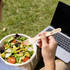 A person holding a wooden fork over a salad bowl, with a laptop in the background on grass. The fork has a logo.