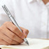 A silver pen held in a hand, poised above a blank notebook on a table.