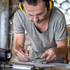 A person wearing safety goggles and ear protection works with tools on a blueprint. The workspace is cluttered with equipment.