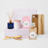 A wooden cheese board, two jars of fudge and coconut ice, a candle, and a diffuser on pink boxes.