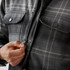 A men's plaid jacket in shades of grey, featuring a Sherpa lining and front pockets, with a zipper being fastened.