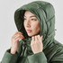 A woman wearing a green puffer jacket with a hood, adjusting it around her face. The jacket has a subtle logo.