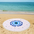 A round beach towel in light pink, featuring an eye design and fringe edges, with a logo present.