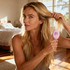 A pink Mini Paddle Brush held by a woman with long, wavy hair in a bright, sunlit bedroom.