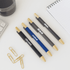 A selection of bamboo click pens in black, blue, and grey, with a notebook and paperclips in the background.